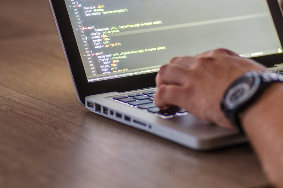 A person using a laptop on a wooden surface, focused on a screen displaying lines of HTML code with highlighted tags and attributes. The individual wears a wristwatch and their hand is positioned over